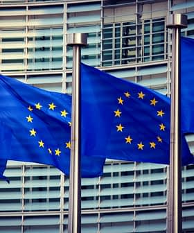 Four European Union flags displayed on poles in front of a modern building. - Olive Oil Times