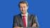 Man wearing a gray suit and a red tie, standing with arms crossed against a blue background. - Olive Oil Times