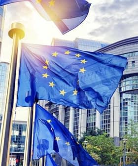 European Union flags waving in front of a modern building with glass architecture. - Olive Oil Times