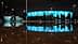 Group of people walking past a modern building with illuminated blue facade at night, reflected in a puddle. - Olive Oil Times