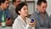 A woman with short hair holding a blue container while seated among a group of people. - Olive Oil Times
