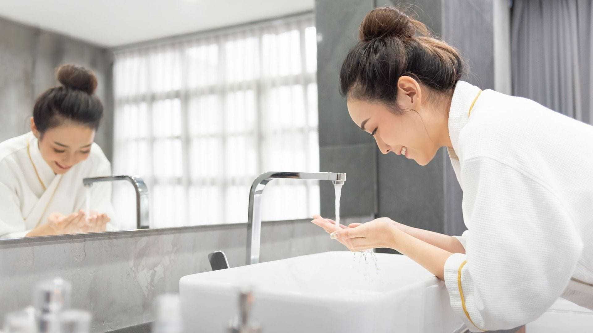 A woman in a white robe washing her hands at a modern sink with flowing water. - Olive Oil Times