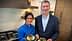 Two individuals, one in a blue chef jacket and the other in a blazer, holding plates of food in a kitchen. - Olive Oil Times